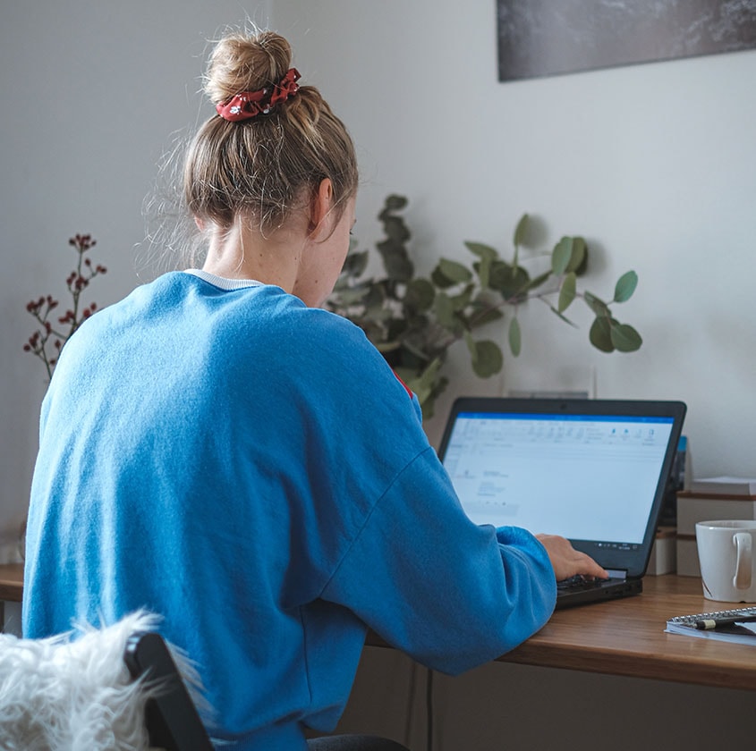 Jeune femme dos à la caméra, vêtu d'un chandail bleu et assise à un bureau. Elle est devant un ordinateur portable.